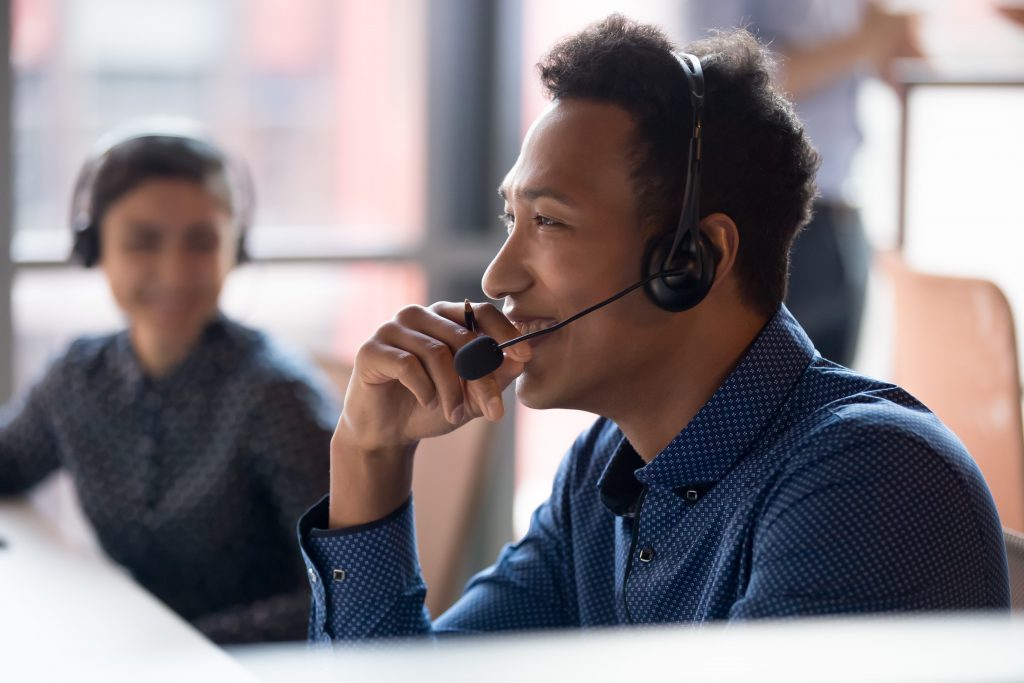 shutterstock_1443614_cKr5r A male adult wearing a headset and smart clothes smiles at something off camera with other adults in the background.