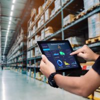 shutterstock_1714734_l3hSi Hands grasp an iPad with a Warehouse Management dashboard on the screen. Cardboard boxes in a warehouse appear behind.