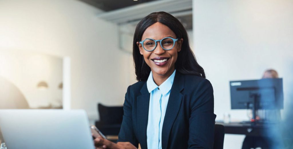 woman-in-office-1024x520 A woman wearing glasses and a suit smiles as the camera as she sits in front of a laptop on a desk holding a mobile phone.