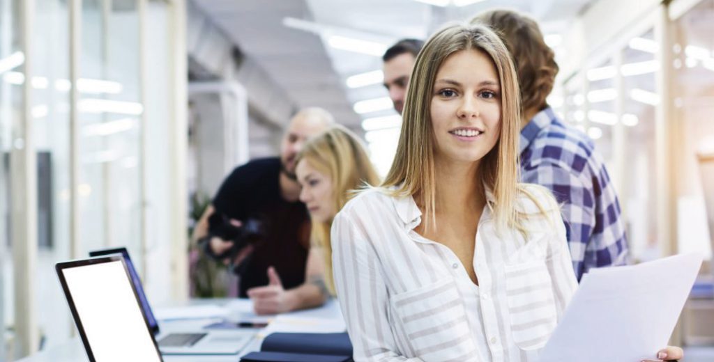 woman-in-office-with-laptop-1024x520 A woman leaning against a desk with a laptop on smiles at the camera as a group of people talk in the background.