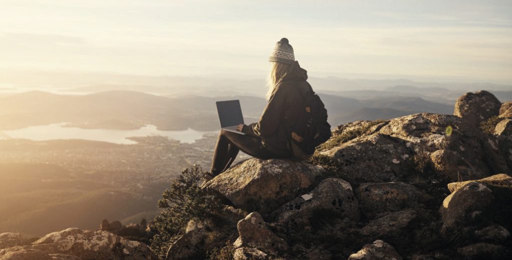 woman_mountain_face-left-1024x520 A woman wearing a bobble hat sits on rocks at the top of mountains with a laptop across her knees, looking out at the view below.