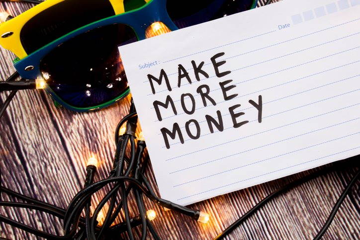 A handwritten note reading 'Make More Money' lies on a wooden surface above fairy lights and sunglasses.